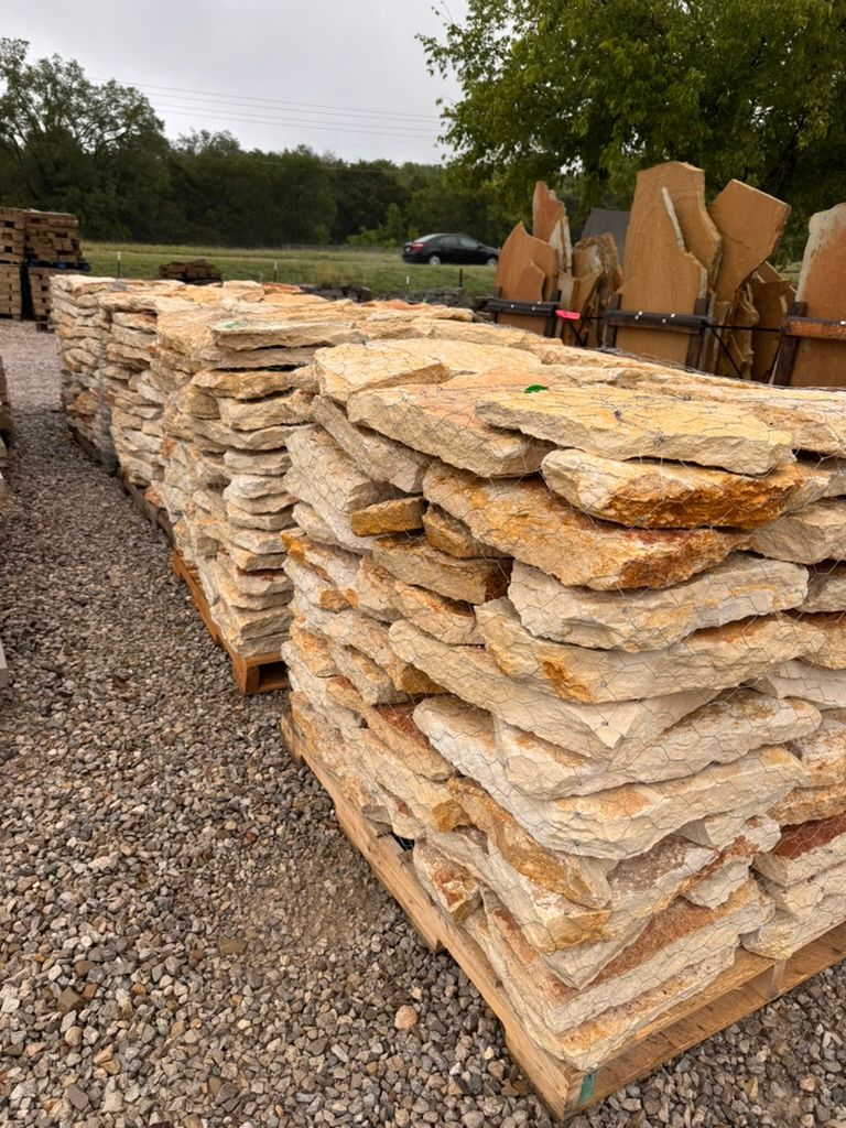 Stacked tan stone slabs on pallets beside a gravel lot, with trees and trucks in the background