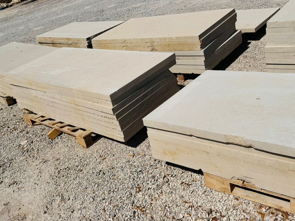 Stacks of large beige stone slabs on wooden pallets in a gravel yard.