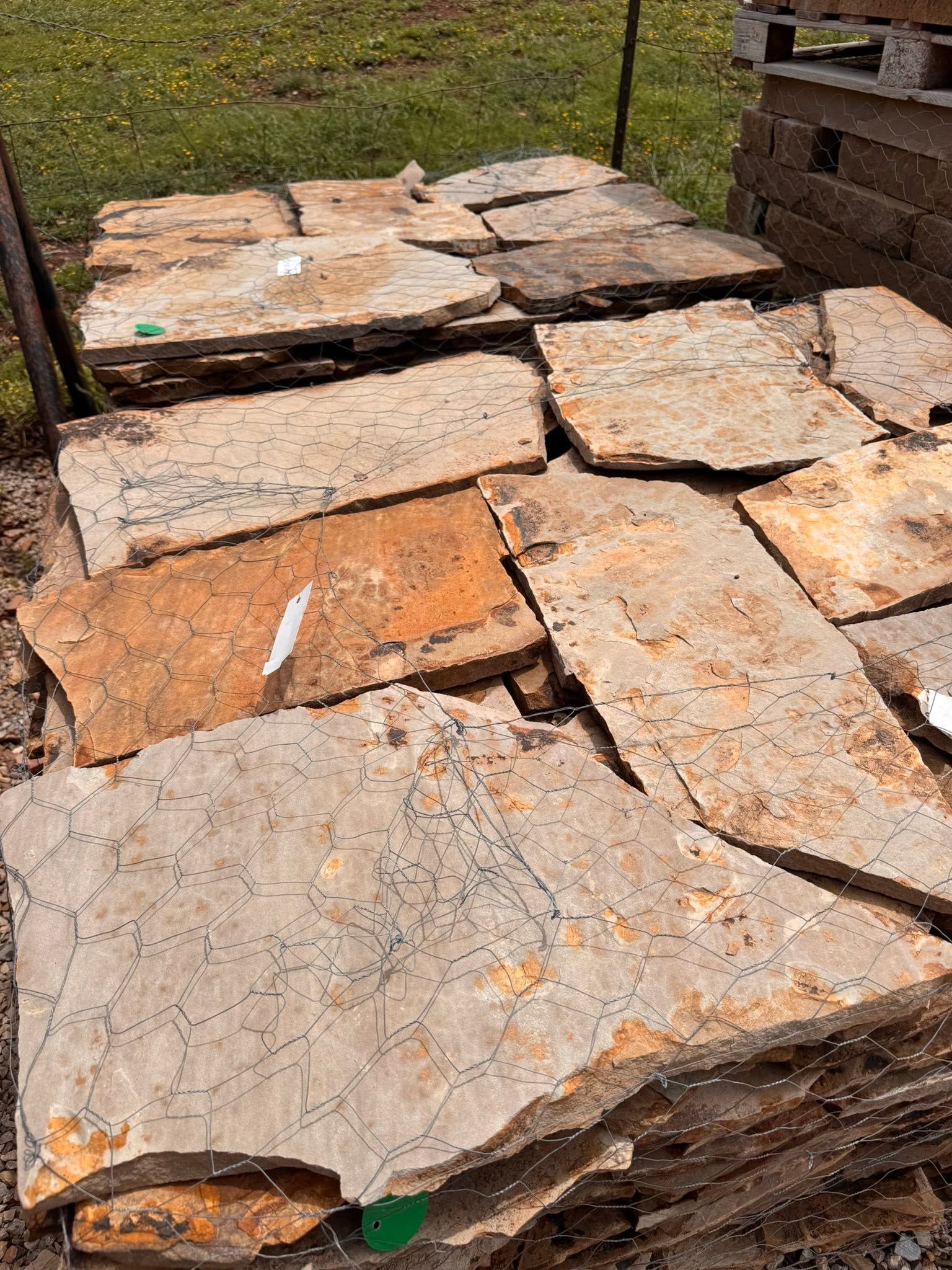 Stacked tan stone slabs outdoors on grass, with a wooden structure in the background.