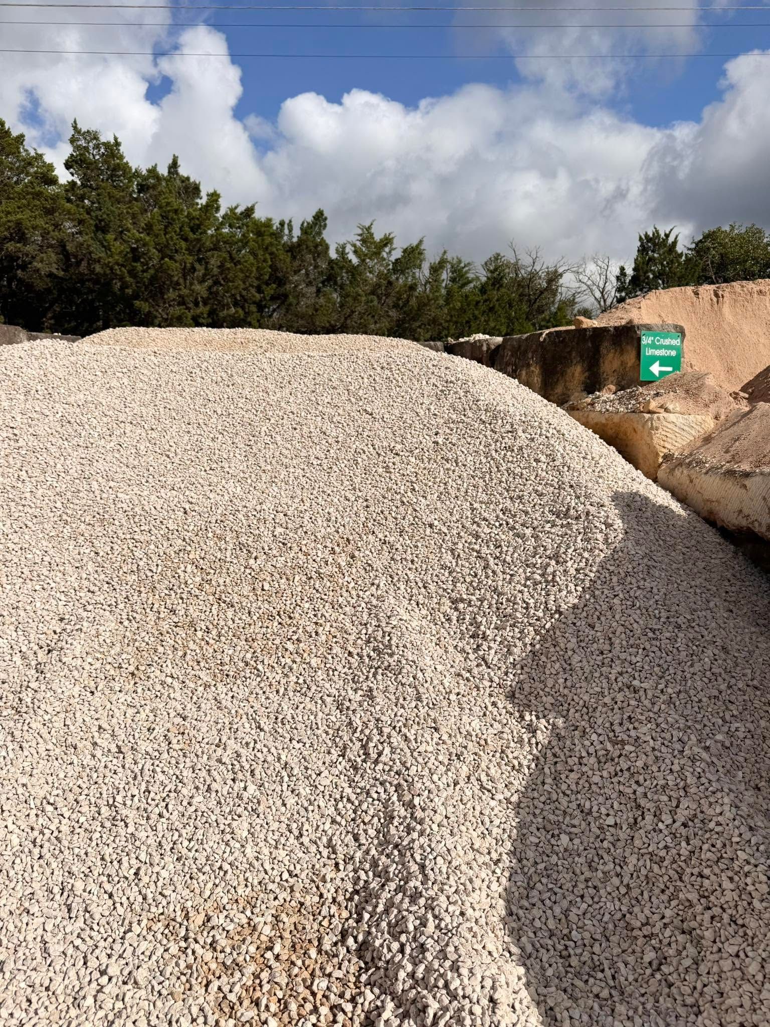 Large pile of light-colored gravel outdoors under a partly cloudy sky, with trees and a small green sign nearby.