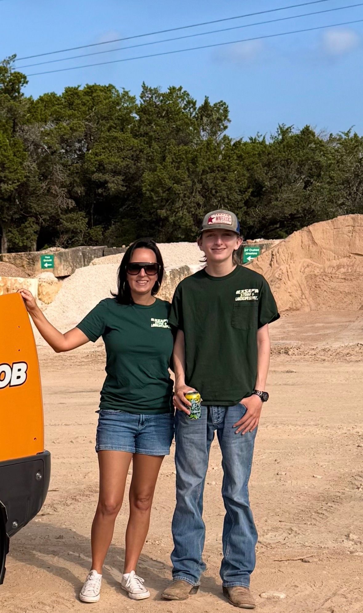 Two people in green shirts and jeans standing outdoors beside an orange object, smiling at the camera.
