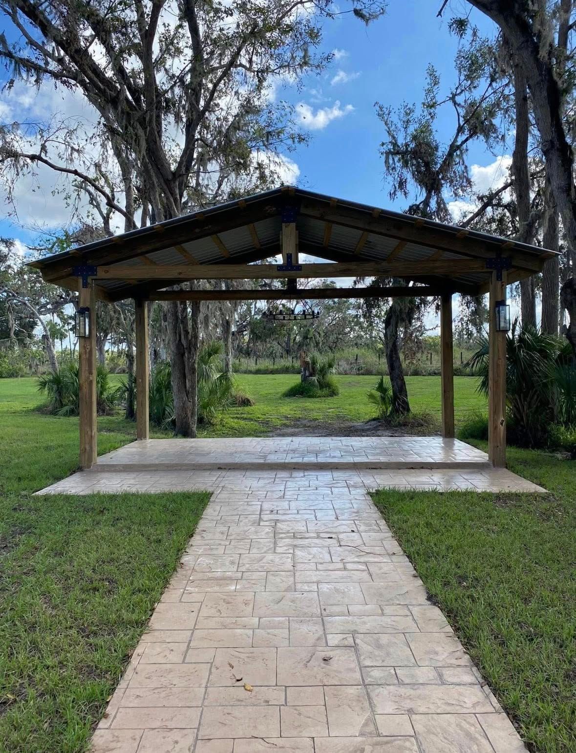 Pavilion with a stamped concrete path, surrounded by green grass and trees. Blue sky visible.