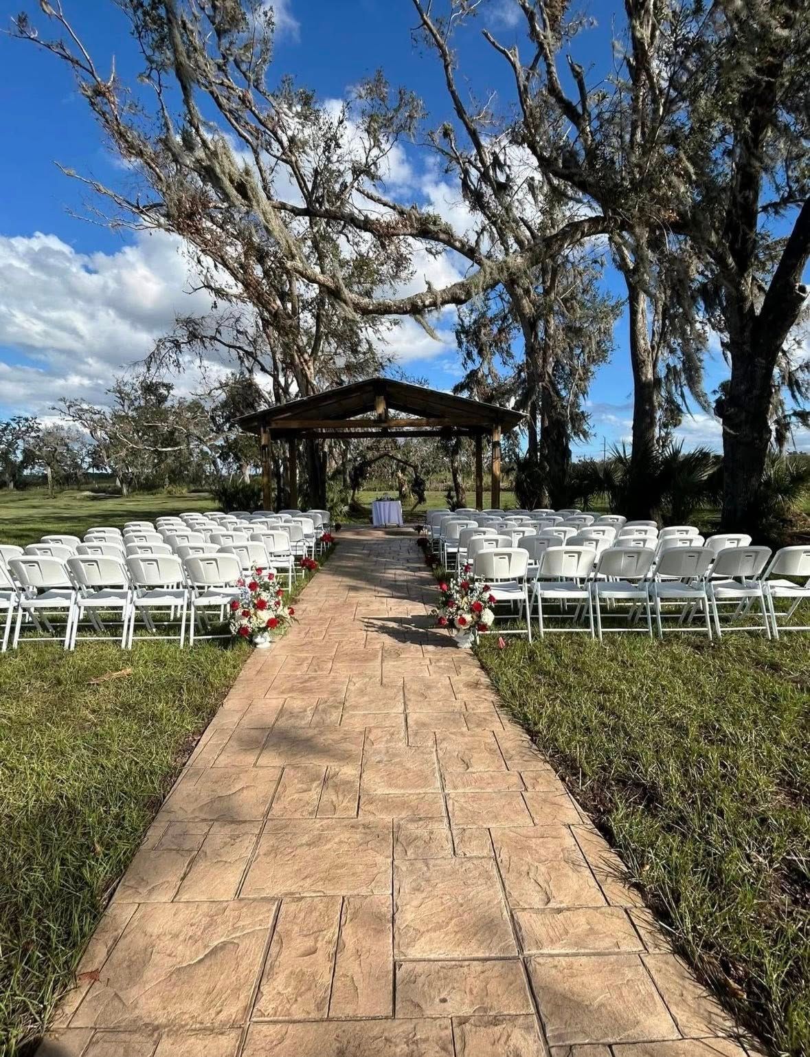 Wedding ceremony setup with white chairs lining a stone path to a gazebo under trees.