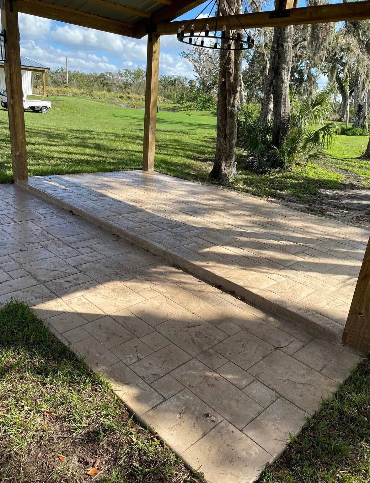 Concrete patio with textured surface under wooden awning; surrounded by green grass and trees.