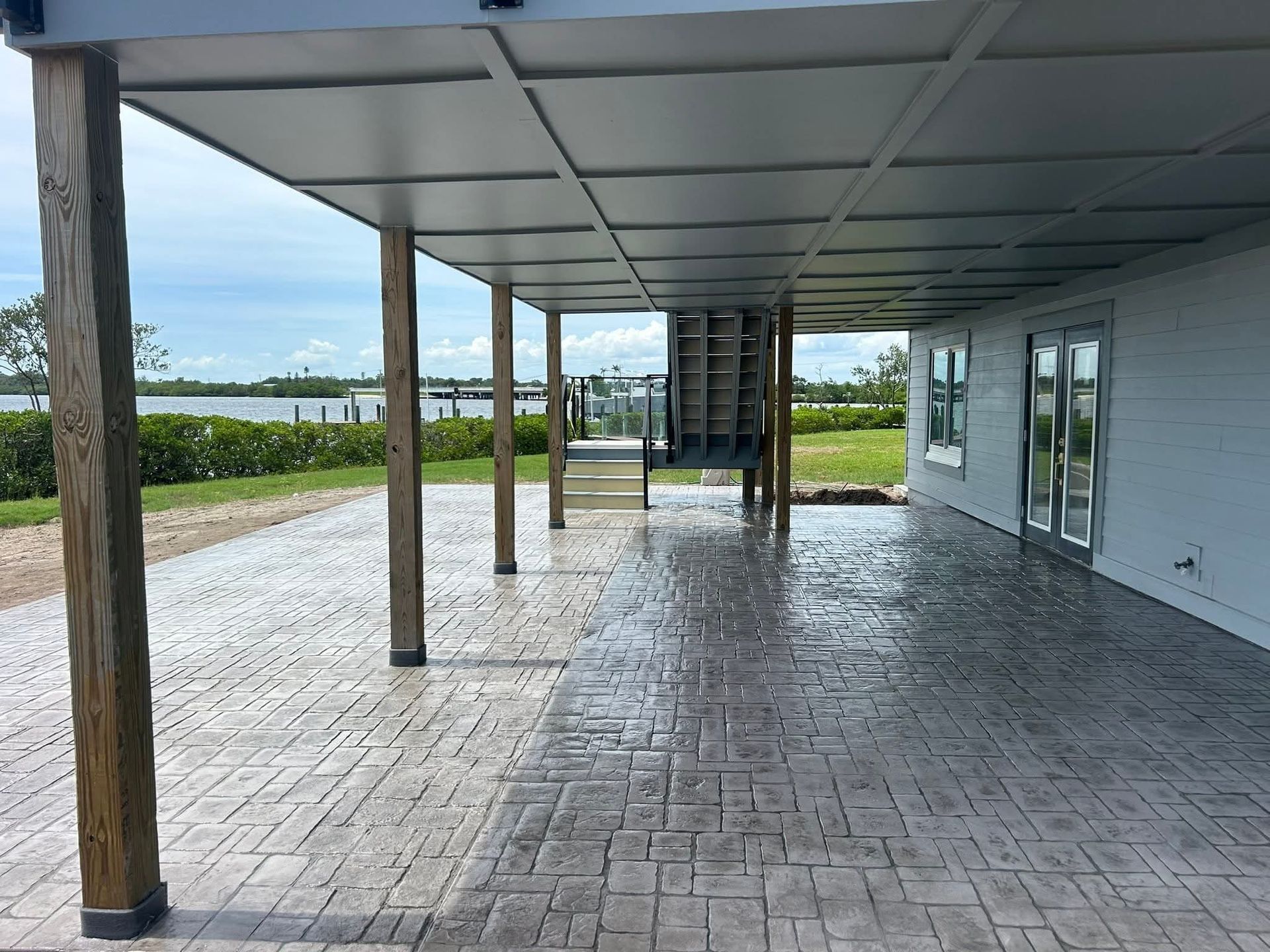Covered patio with stamped concrete flooring, wooden posts, and a view of water and a building.