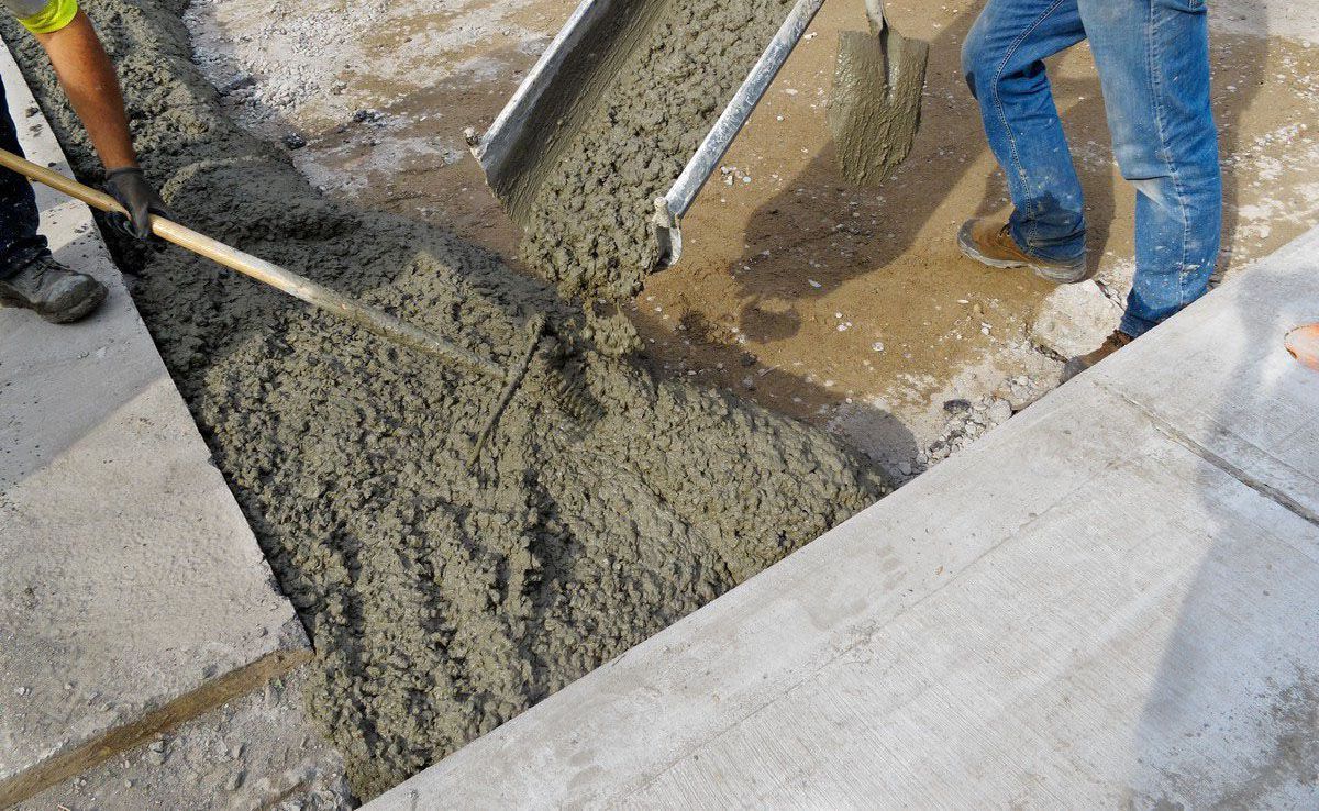 Workers pouring concrete into a form between existing concrete structures with tools and equipment.
