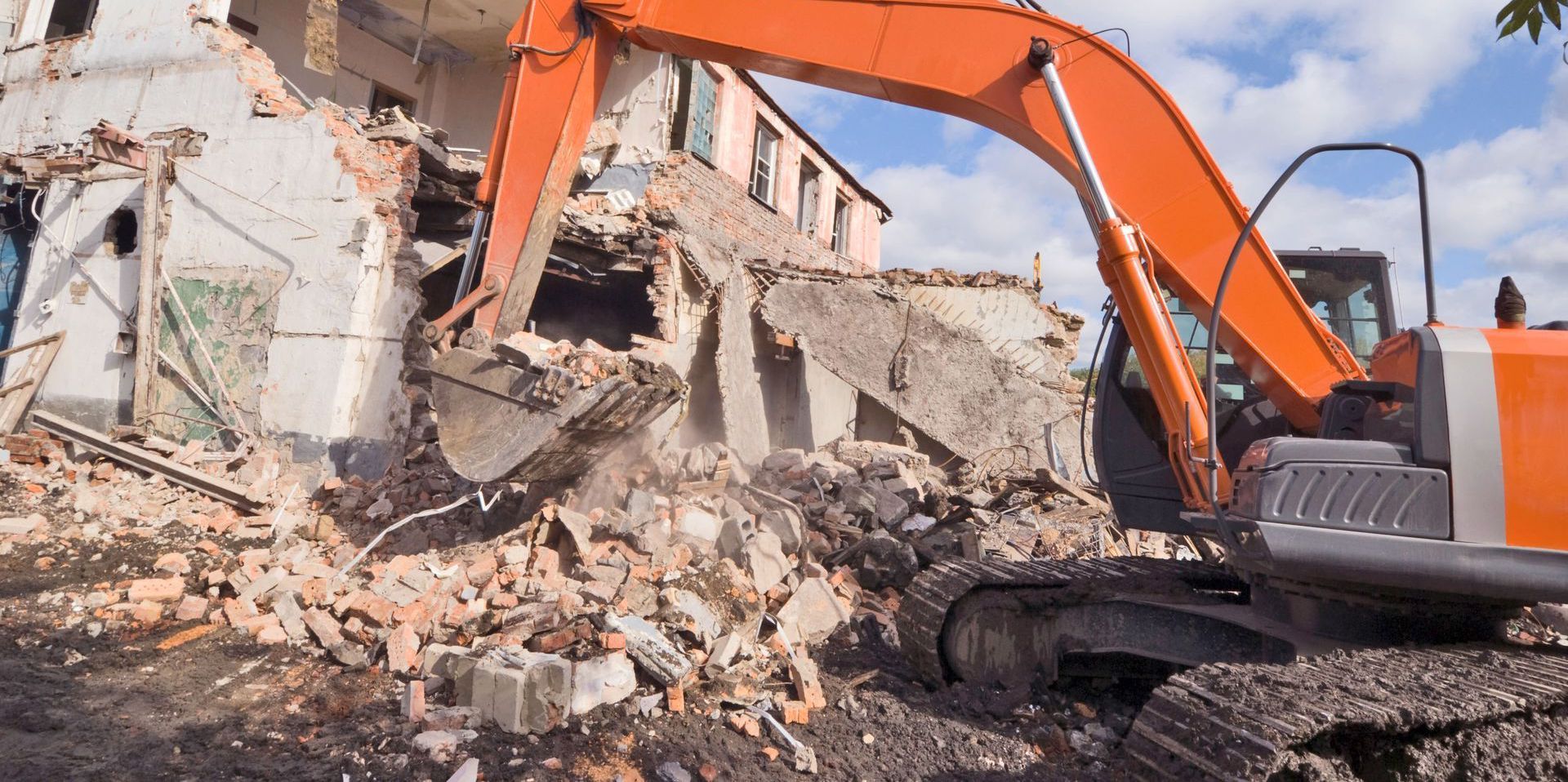 An orange excavator demolishes a building, creating rubble.