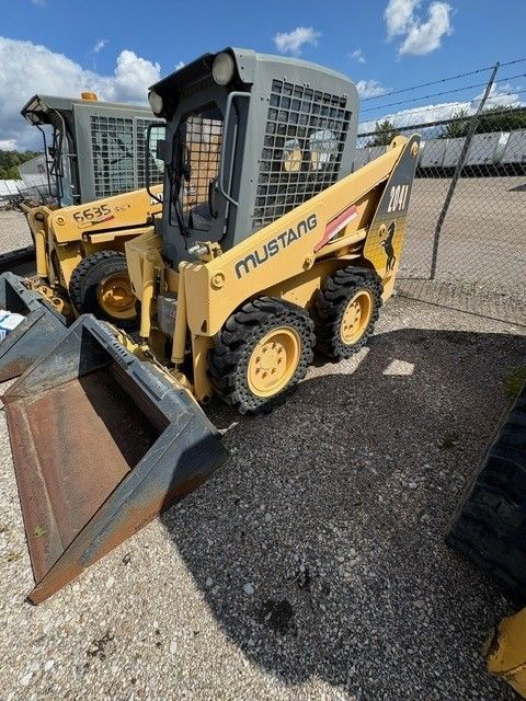 Yellow Mustang skid-steer loader with a bucket in a yard on a sunny day.