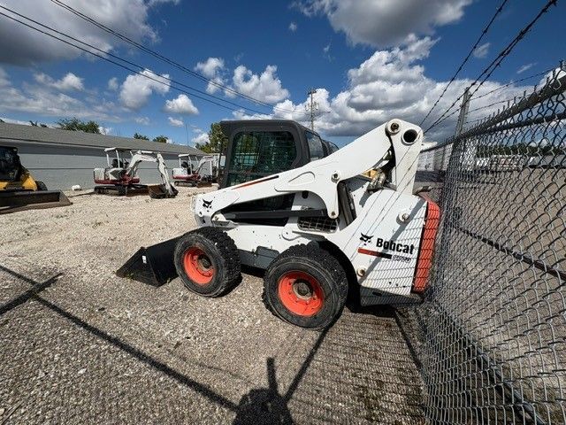 White Bobcat skid-steer loader parked outside, orange wheels, black bucket, chain link fence, blue sky.
