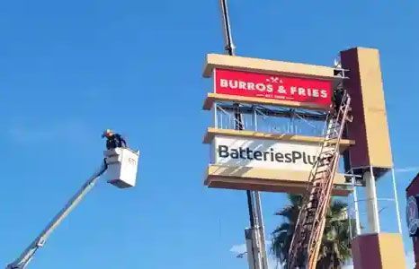 Worker in a bucket lift beside a Burros & Fries and Batteries Plus sign against a blue sky