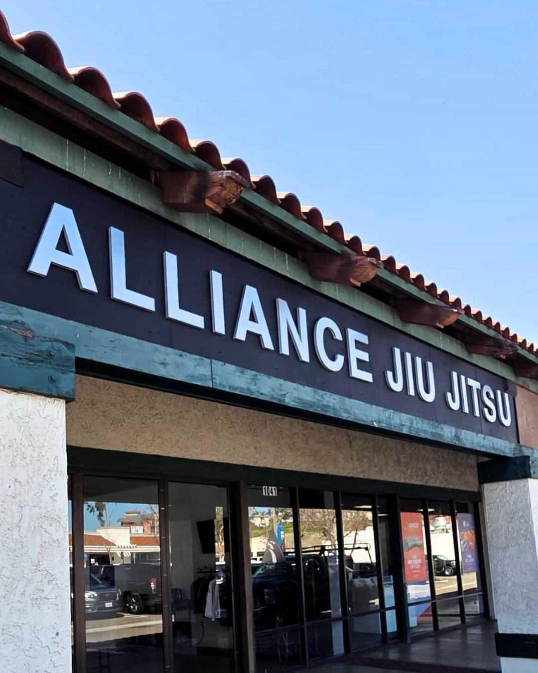 Storefront sign reading Alliance Jiu Jitsu on a strip mall building under a blue sky.