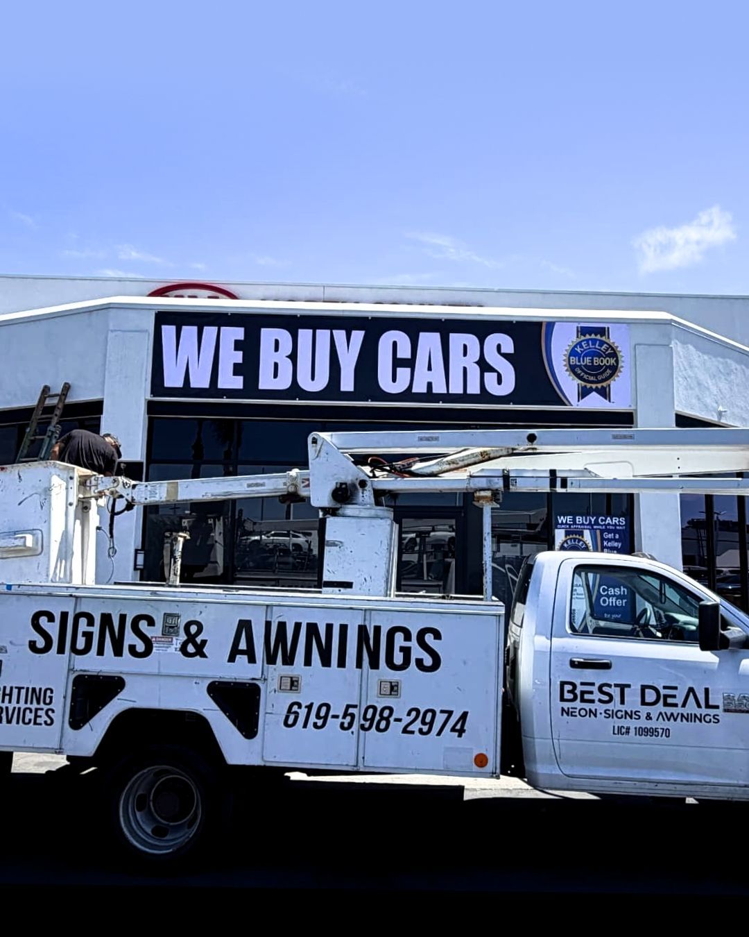 Tow truck with WE BUY CARS and SIGNS & AWNINGS signage parked outside a shop.