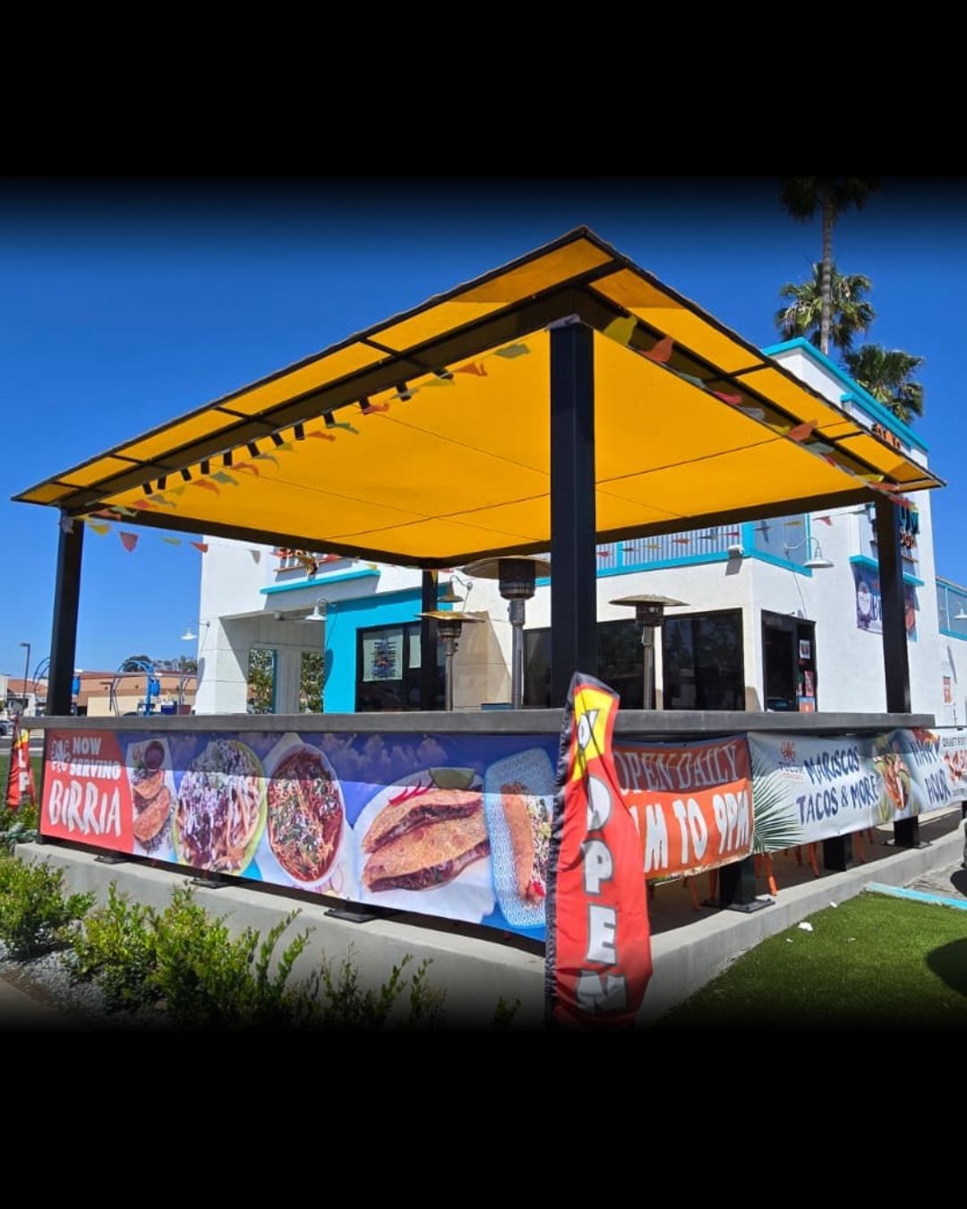Outdoor food kiosk with a bright yellow roof and colorful menu banners on a sunny day.