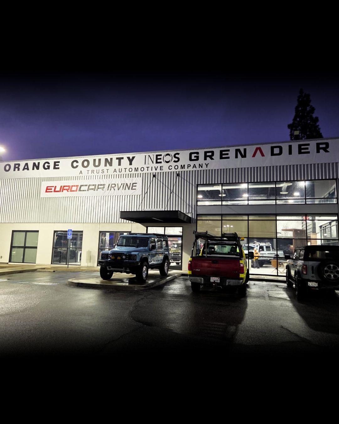 Orange County Jeep Grenadier dealership at night, with parked vehicles and wet pavement reflections