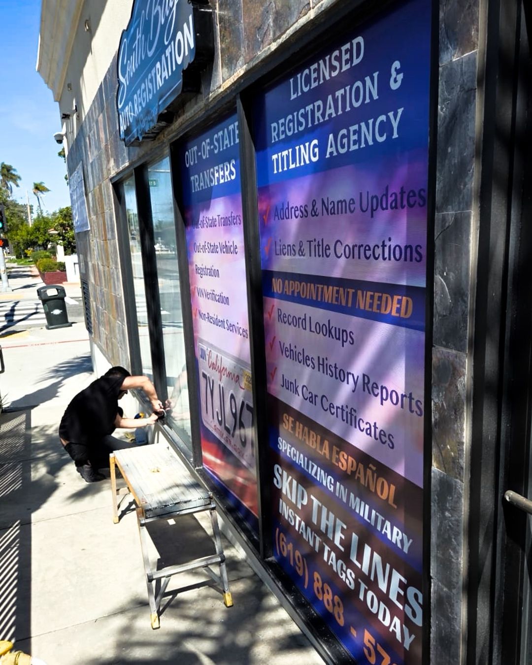A person kneels by a storefront window, painting a sign beside a sidewalk and parked cars.