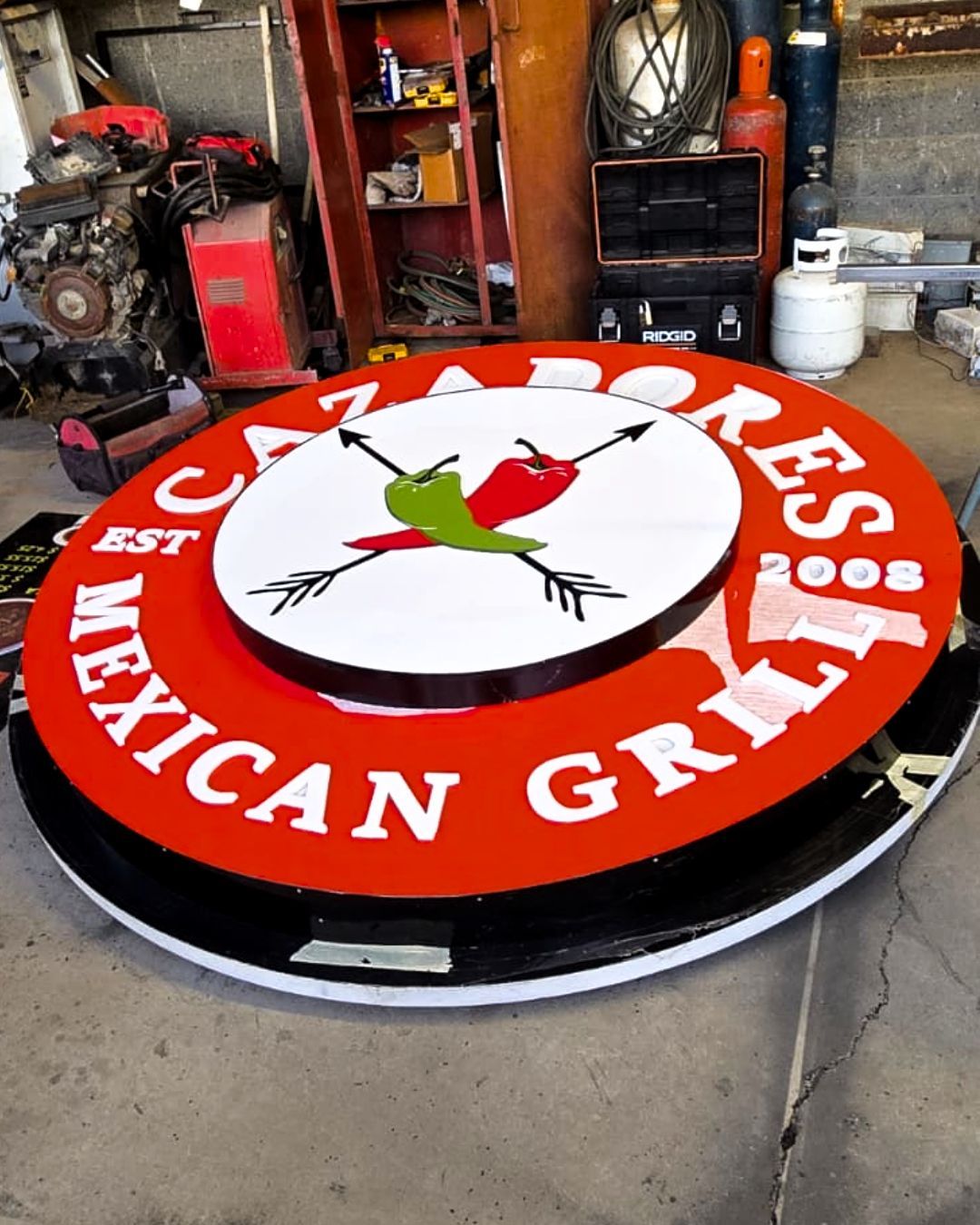 Round red-and-white Los Taqueros Mexican Grill sign with a chili pepper logo in a workshop garage
