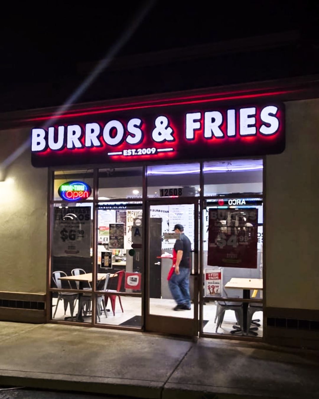 BURROS & FRIES fast-food restaurant storefront at night, with a neon sign and a person inside near the door