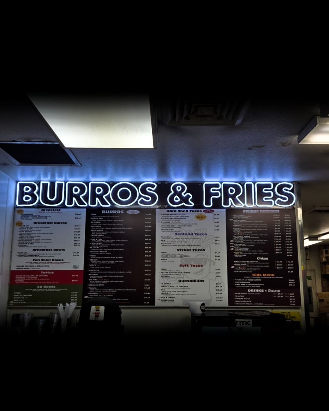 Neon sign reading BURROS & FRIES above a restaurant menu board at night