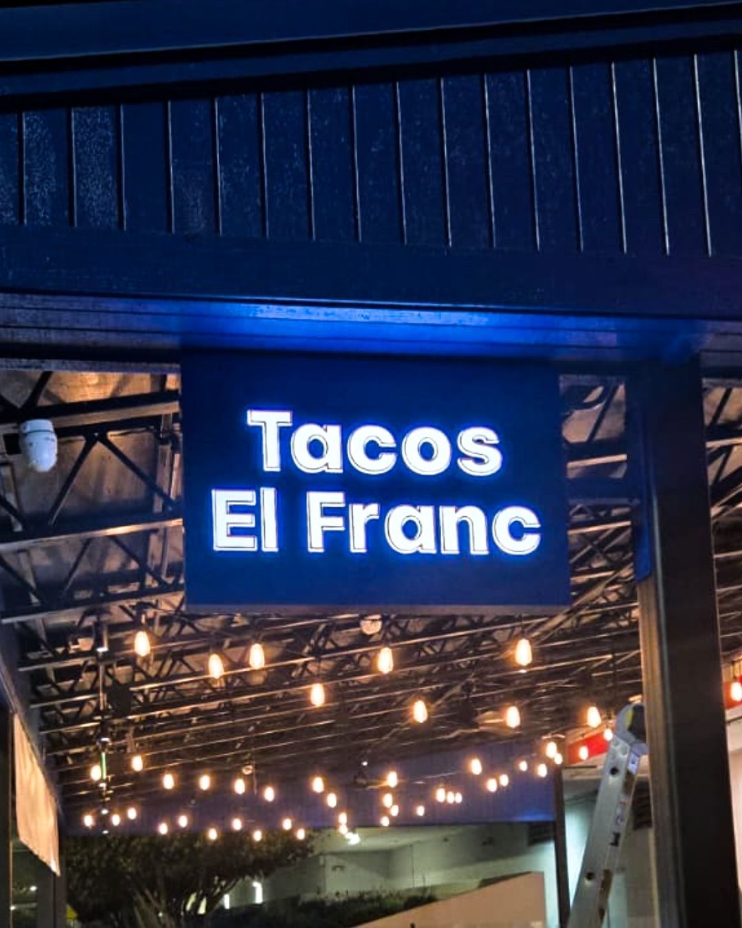 Blue-lit sign reading Tacos El Franc above a restaurant entrance with string lights.