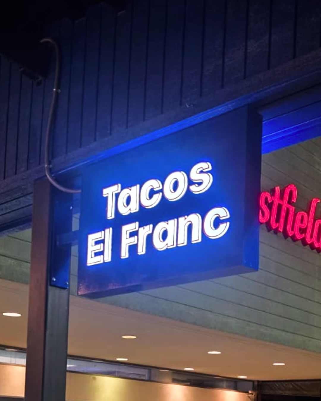Blue illuminated sign for Tacos El Franc outside a storefront at night