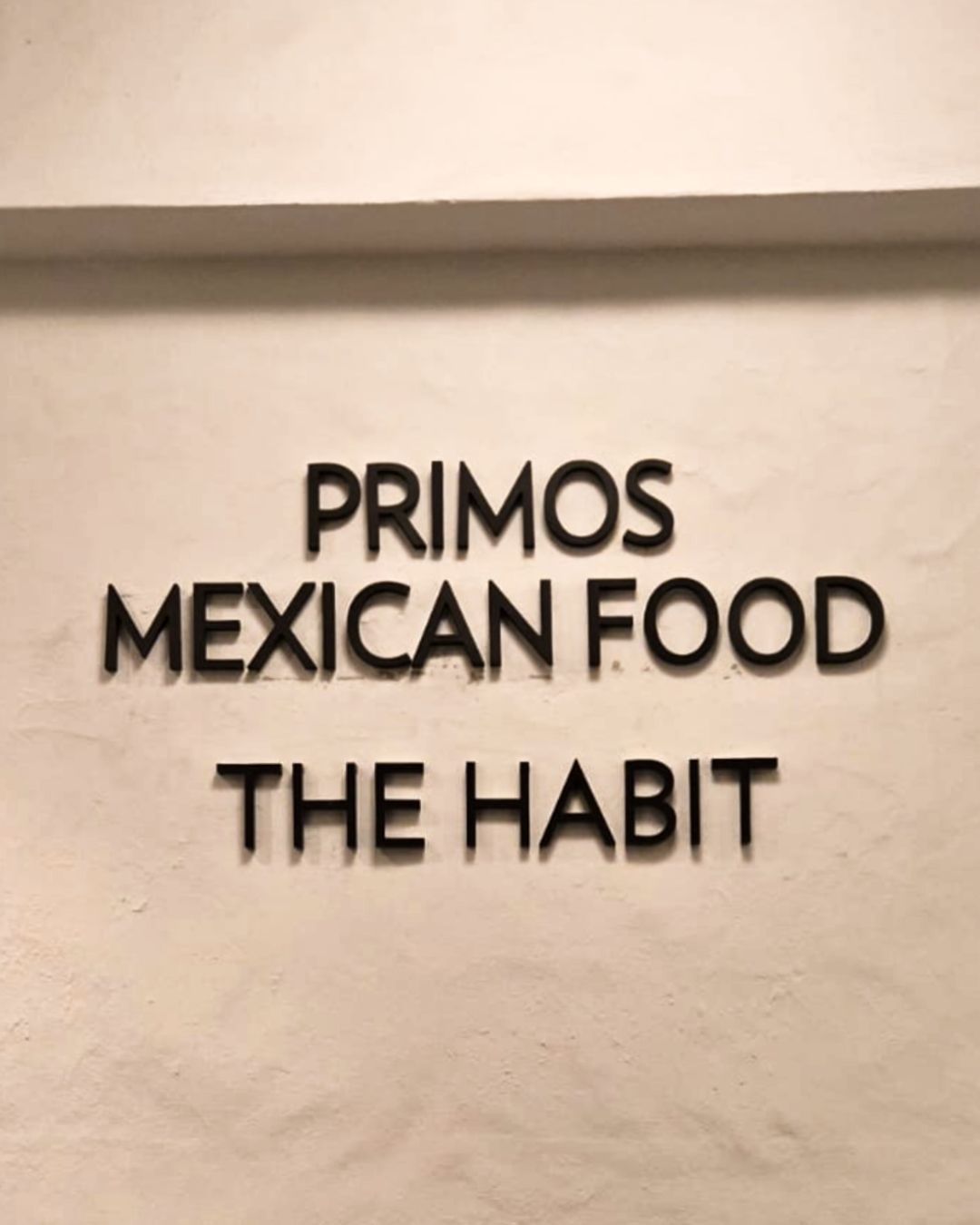 Black wall sign reading PRIMOS MEXICAN FOOD and THE HABIT in raised letters.