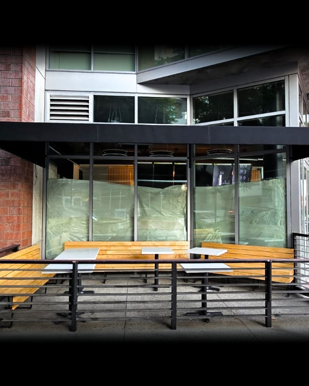 Outdoor café seating under a black awning beside a building, with wooden benches and tables behind glass panels