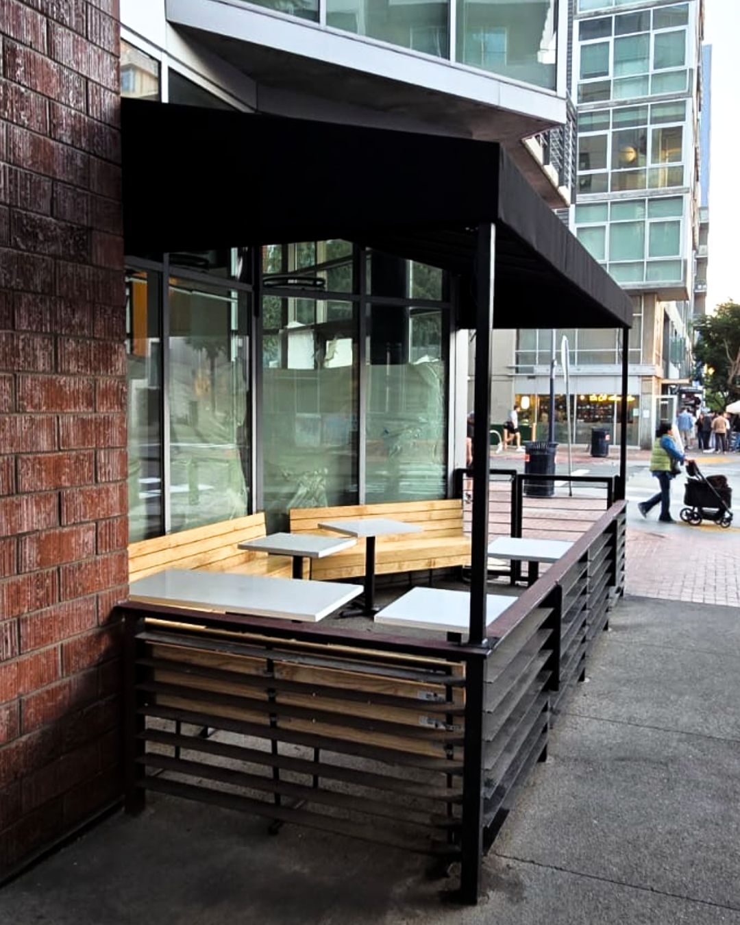 Empty outdoor café patio with wooden benches under a black awning beside a city sidewalk