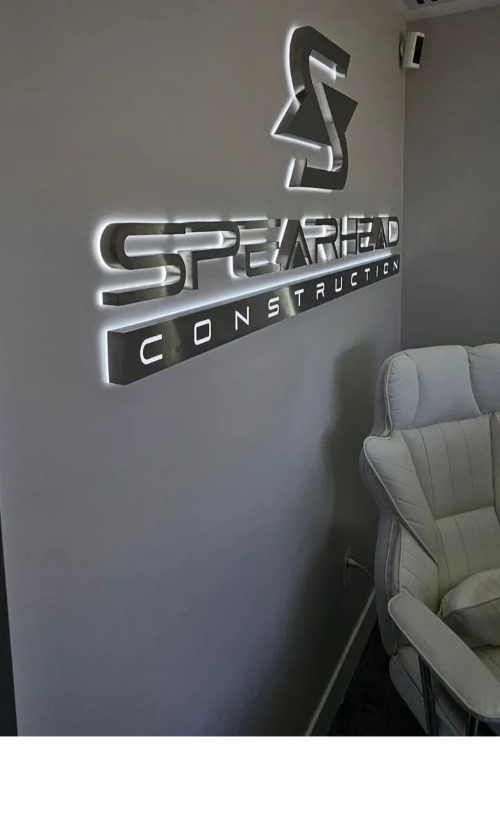 Silver logo sign on a gray wall beside a light-colored chair in an office or lobby