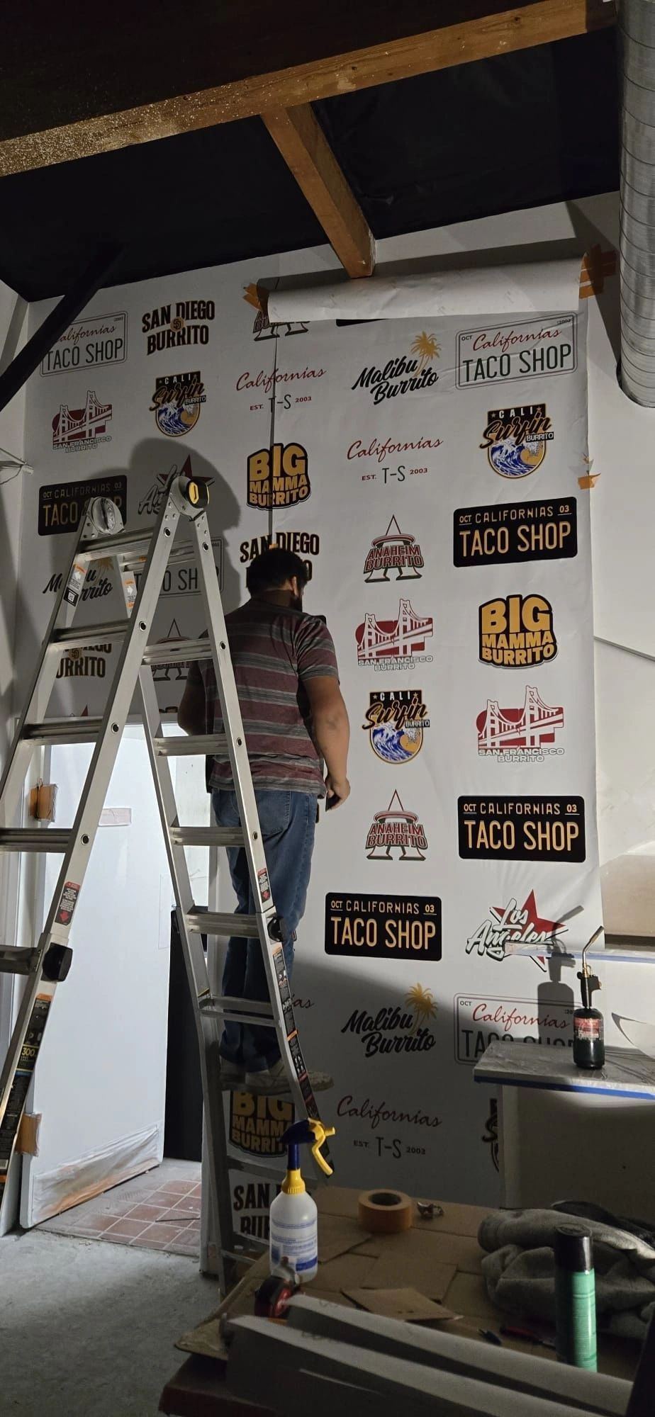 Person using a ladder to hang wall art in a white room with exposed ceiling beams
