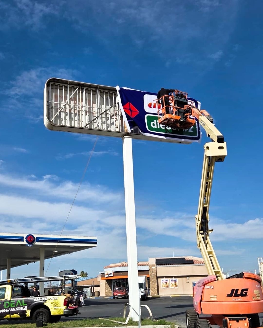 A worker on a lift is cleaning a billboard beside a gas station under a blue sky