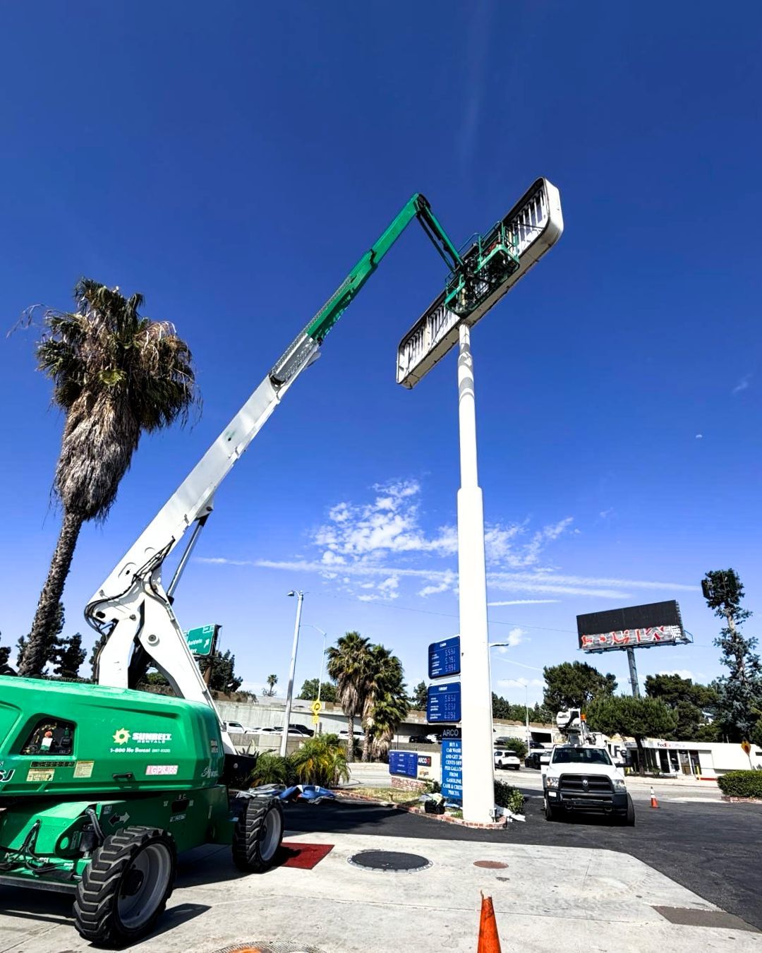 A green boom lift hoists a billboard sign on a sunny roadside with palm trees and parked cars