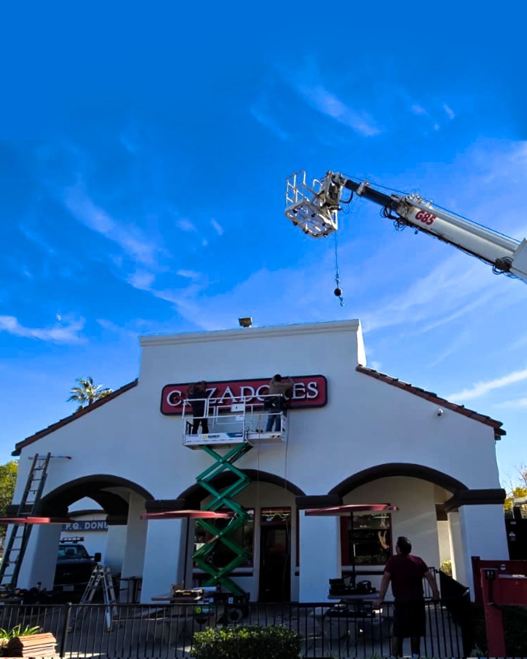 Crane lifting a sign onto a white storefront under a bright blue sky