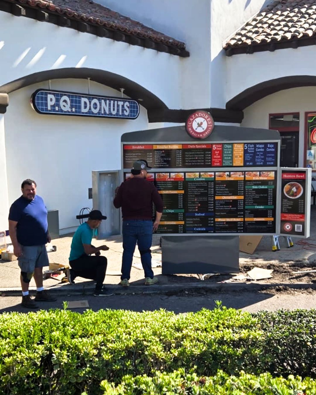 People reading a menu board outside P.Q. Donuts in a sunny courtyard