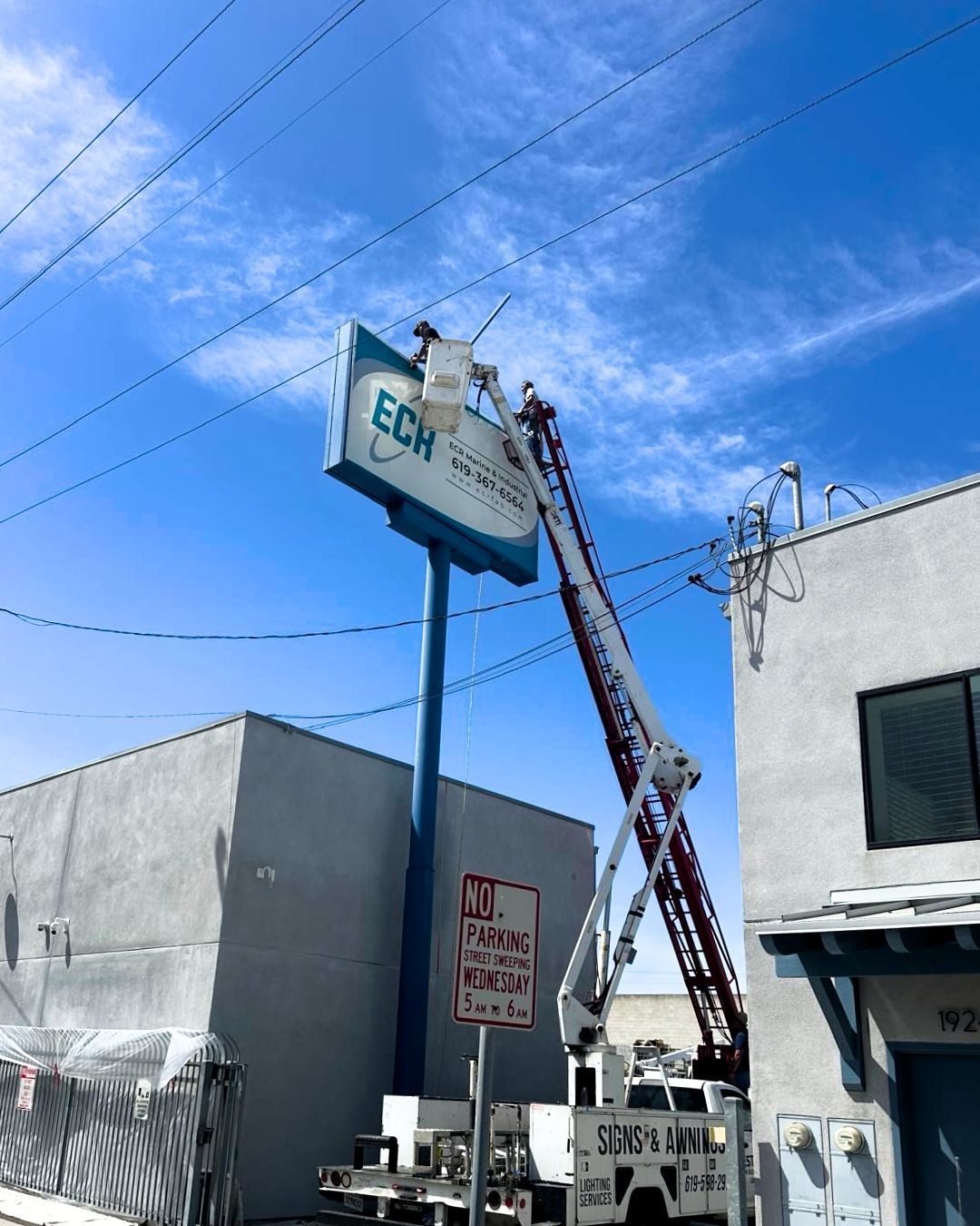 Workers in a bucket lift are repairing a tall sign on a blue pole between buildings under a clear sky
