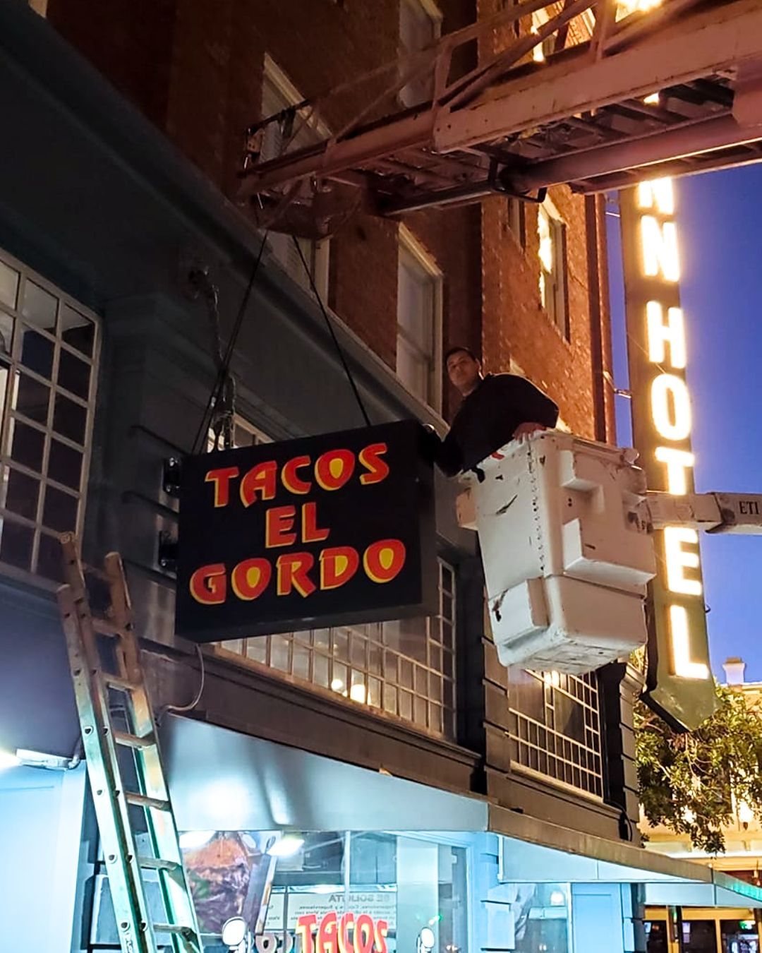 Neon sign for Taco El Gordo on a nighttime street, next to a vertical hotel sign.