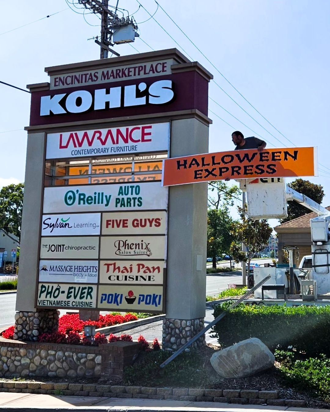 Shopping center sign with Kohl’s, Lawrence, and Halloween Express ads; person atop the sign under a blue sky