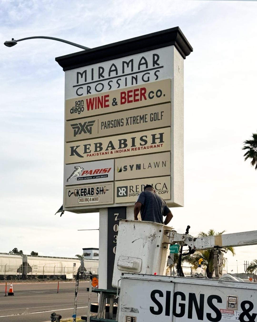 Tall roadside sign for Miramar Crossings with business listings, beside a worker on a lift.