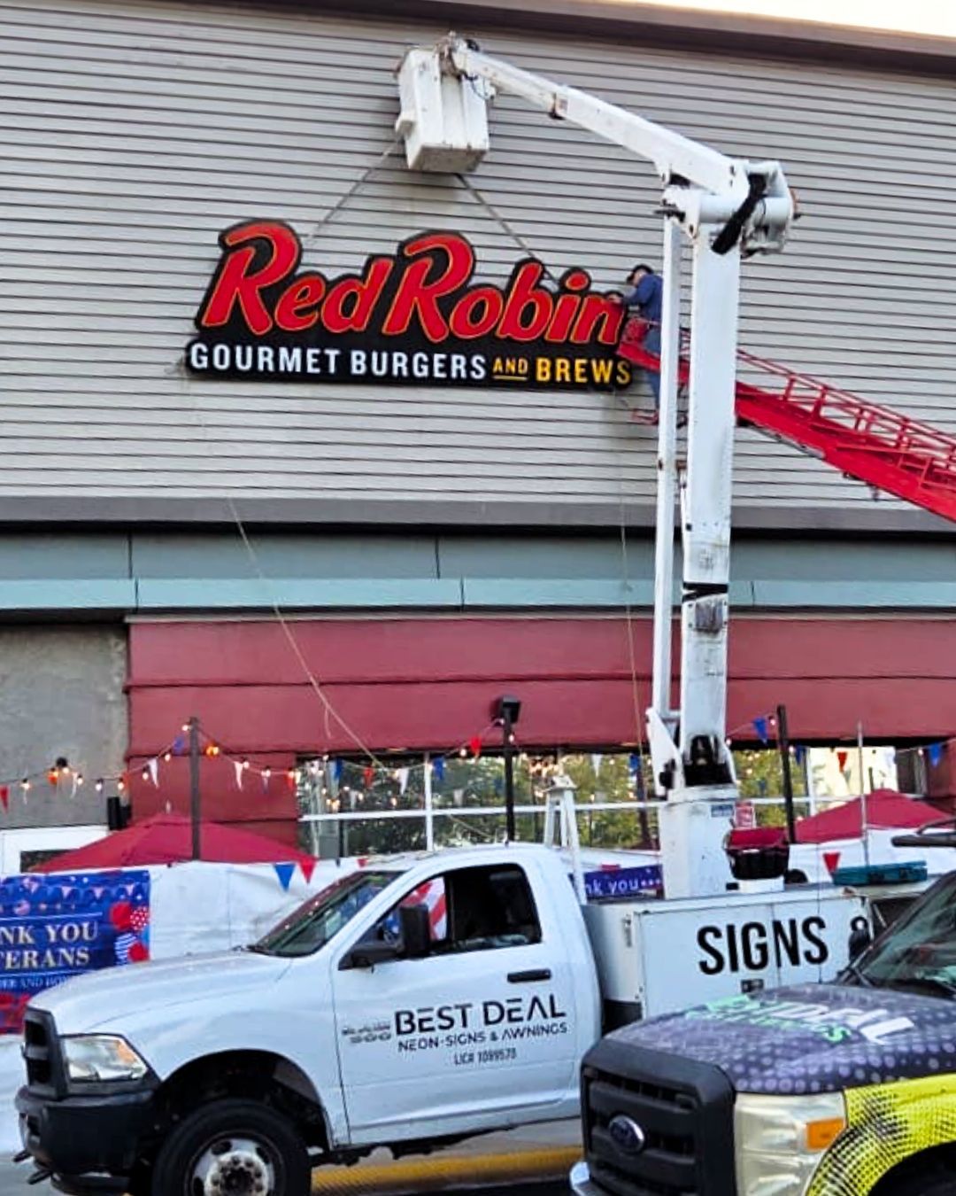 Worker in a bucket lift outside Red Robin restaurant, painting or installing signage near parked trucks.