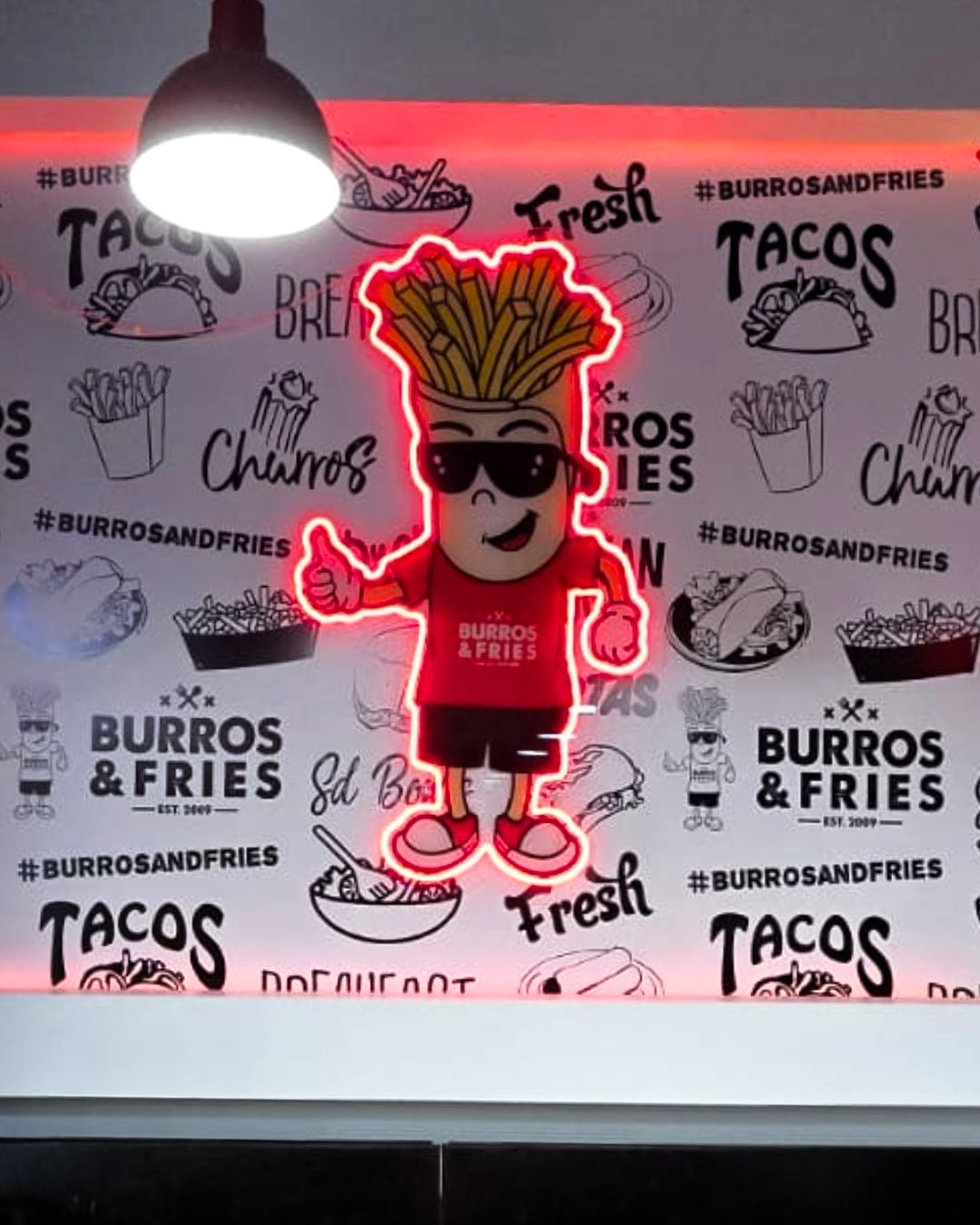 Neon sign of a smiling cartoon fries mascot in sunglasses against a taco and burrito restaurant wall
