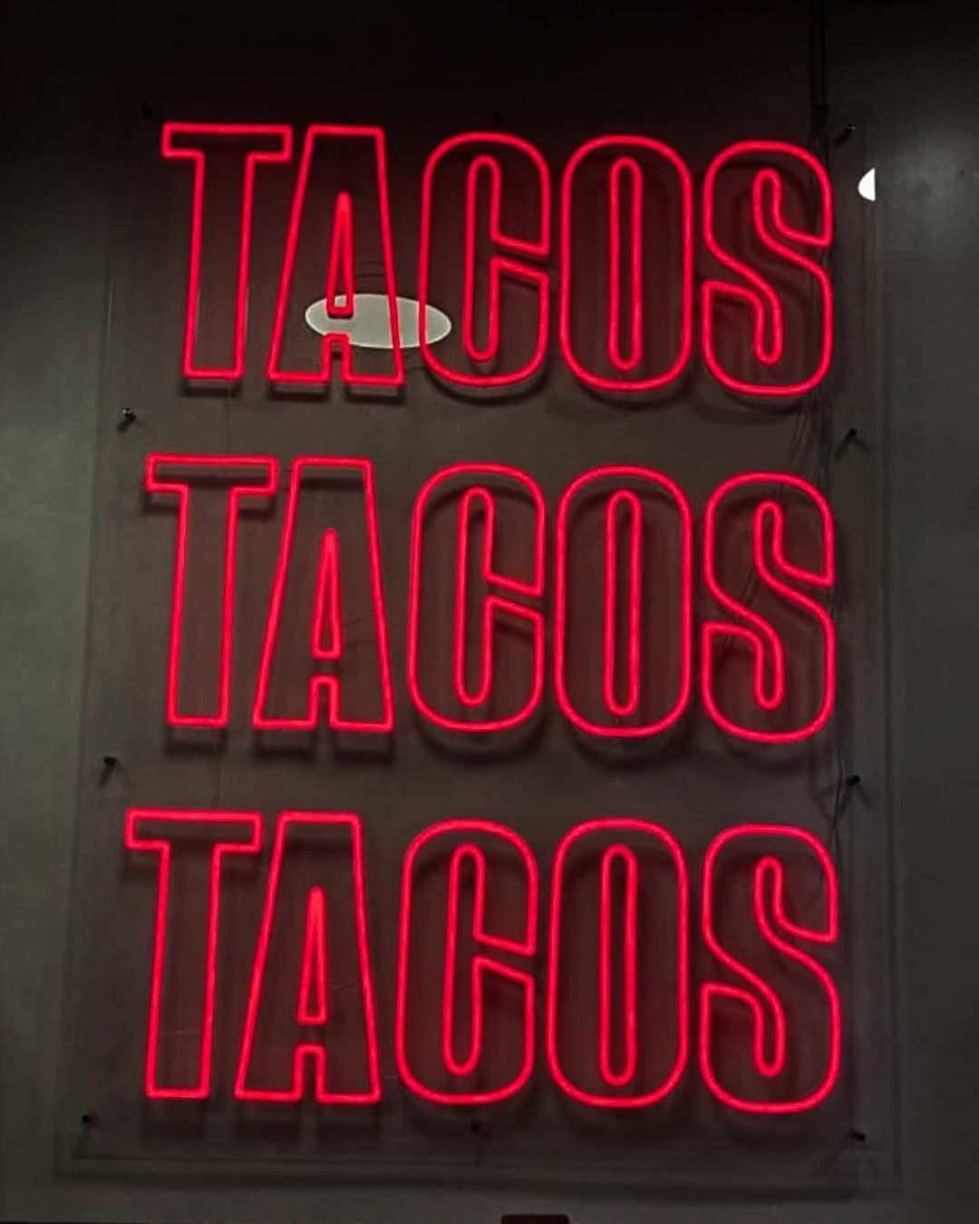 A neon red sign reading TACOS repeated three times on a dark wall.