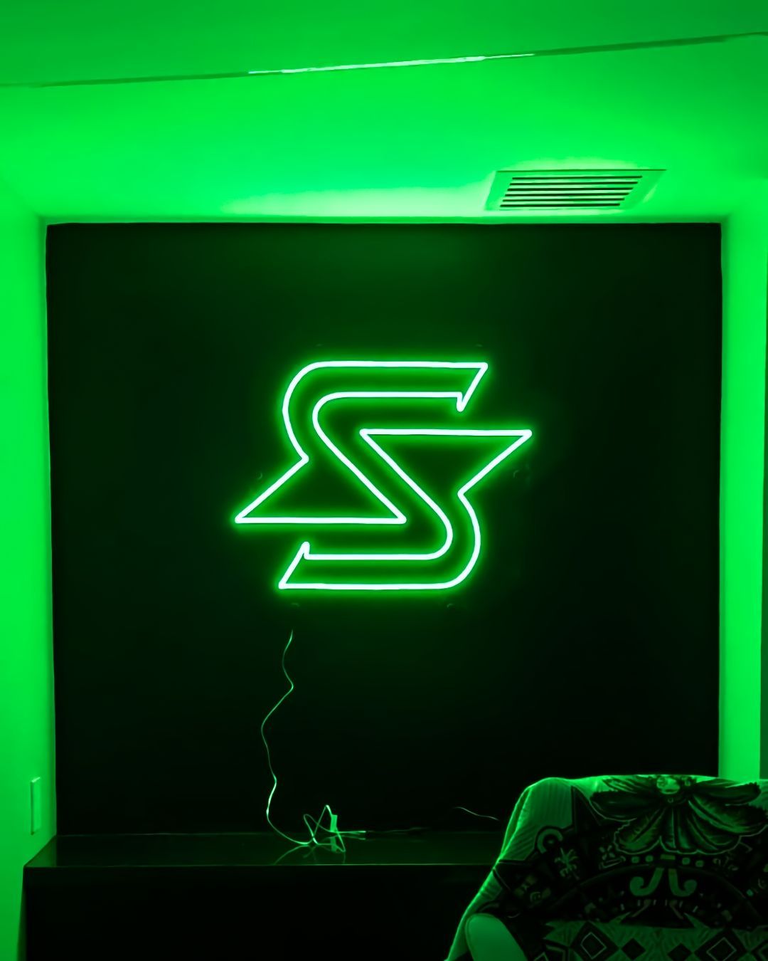 Neon green S logo glowing on a black wall in a dim room with green ambient lighting.