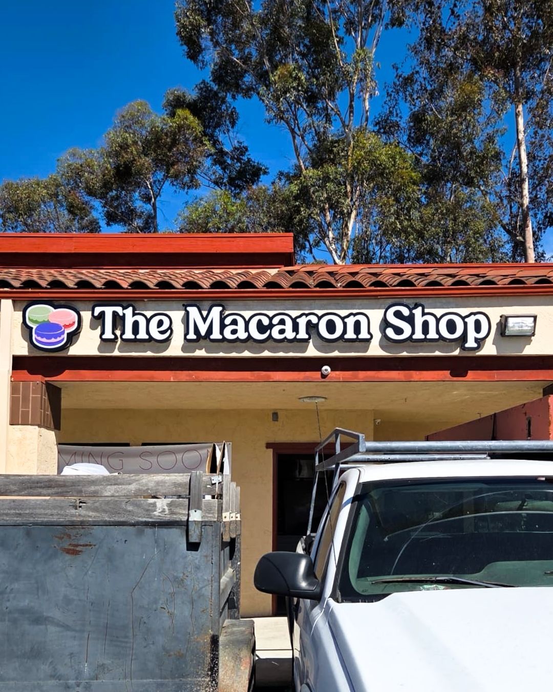 Storefront sign for The Macaron Shop with trees and parked vehicles in front