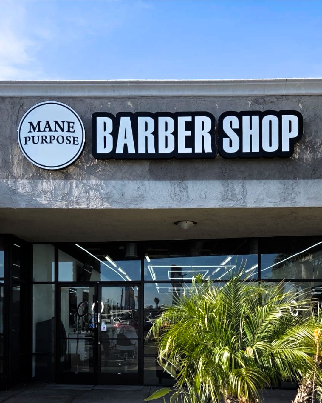 Storefront sign reading BARBER SHOP above a glass entrance with a palm plant in front.