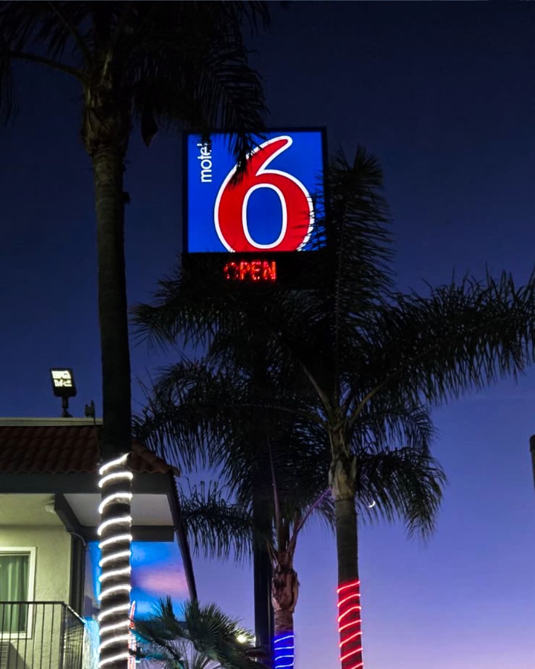 Motel 6 neon sign glowing at dusk above palm trees and a building with lit columns