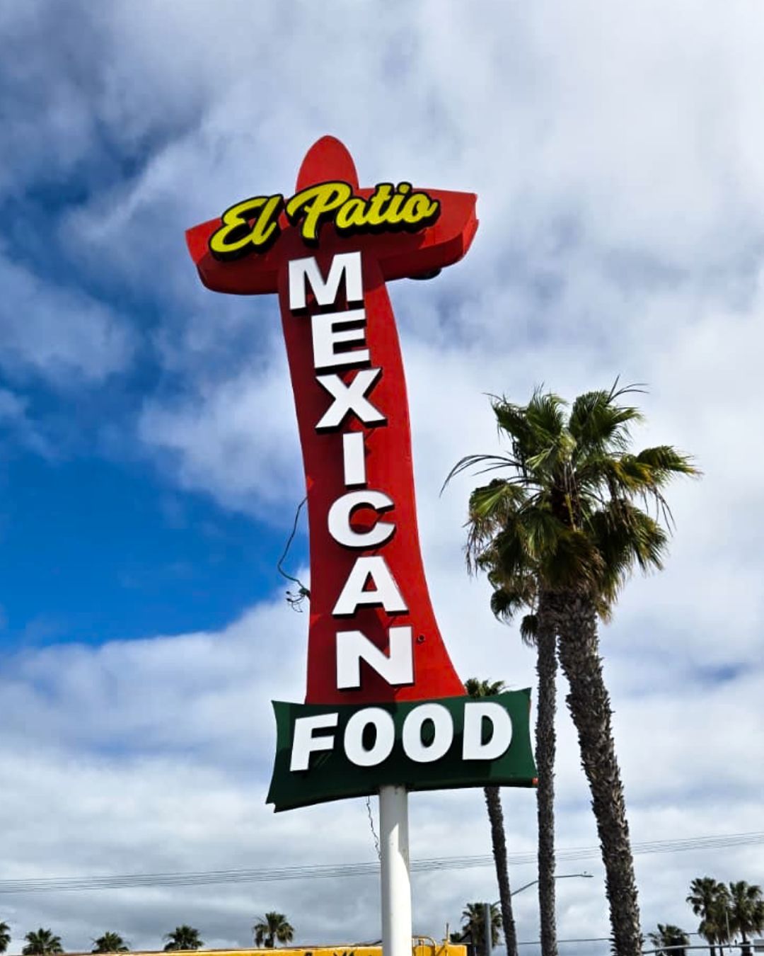 El Patio Mexican Food sign on a tall red pole against a blue sky with palm trees