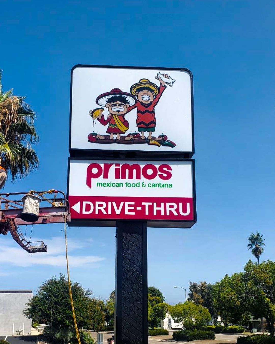 Primos Mexican food sign with cartoon characters and a drive-thru marker against a blue sky