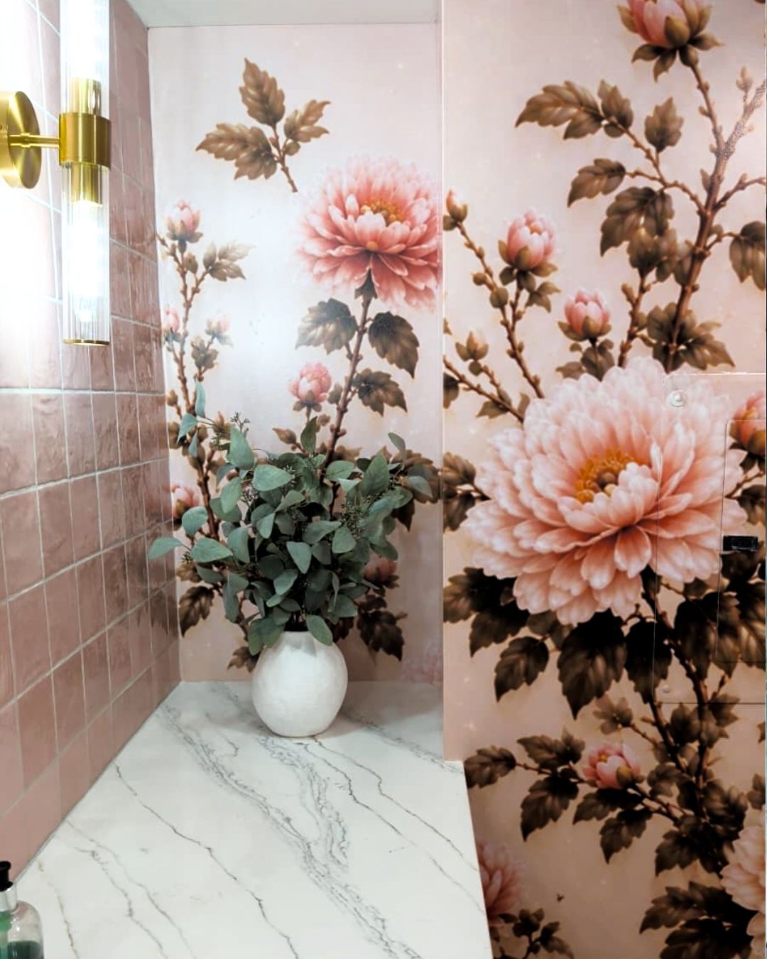 Floral wallpaper in a pink bathroom with a white marble counter and a vase of green leaves and flowers