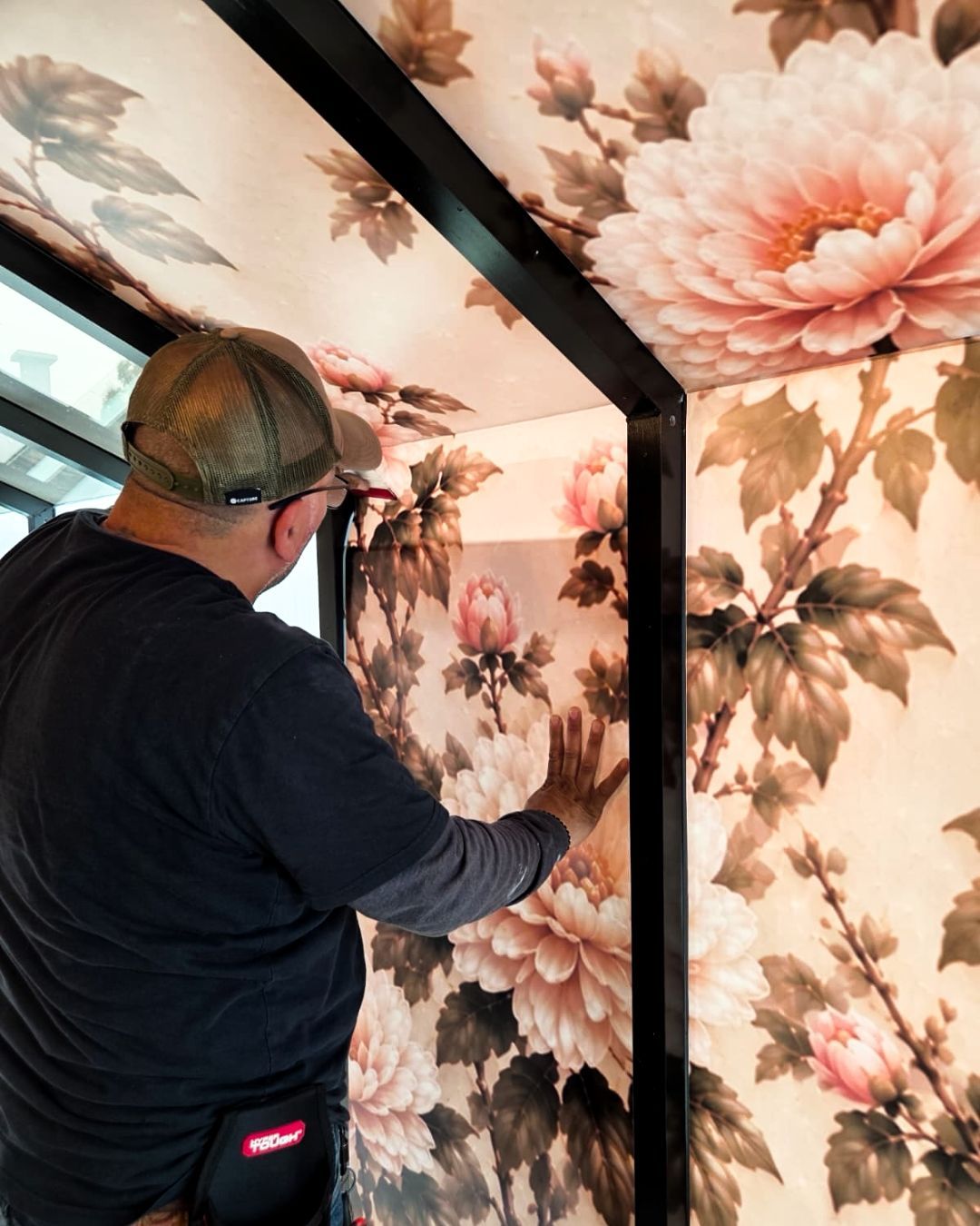 A man hanging floral wallpaper on a wall in a room with black trim