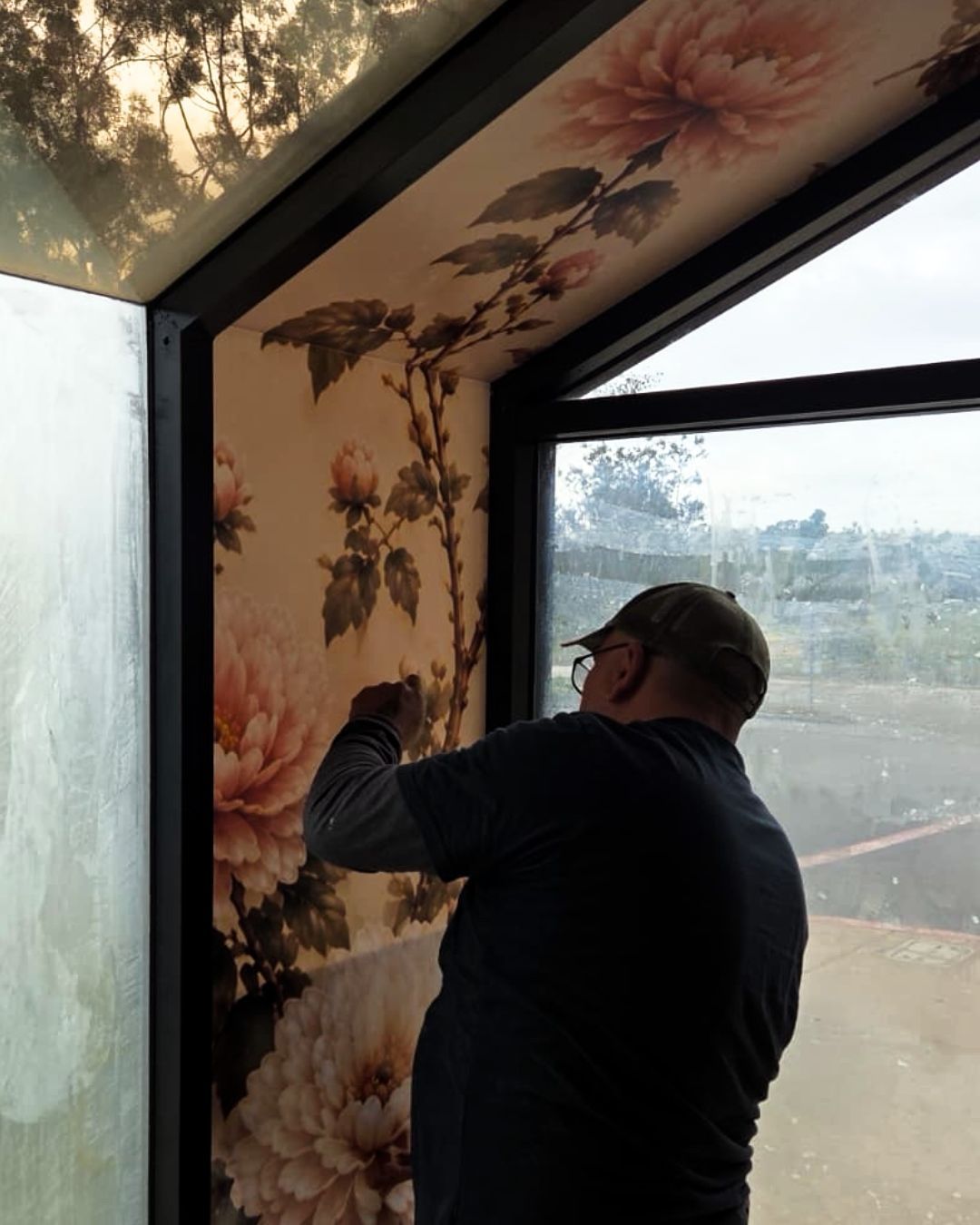 A person applying floral wallpaper inside a room with large windows.