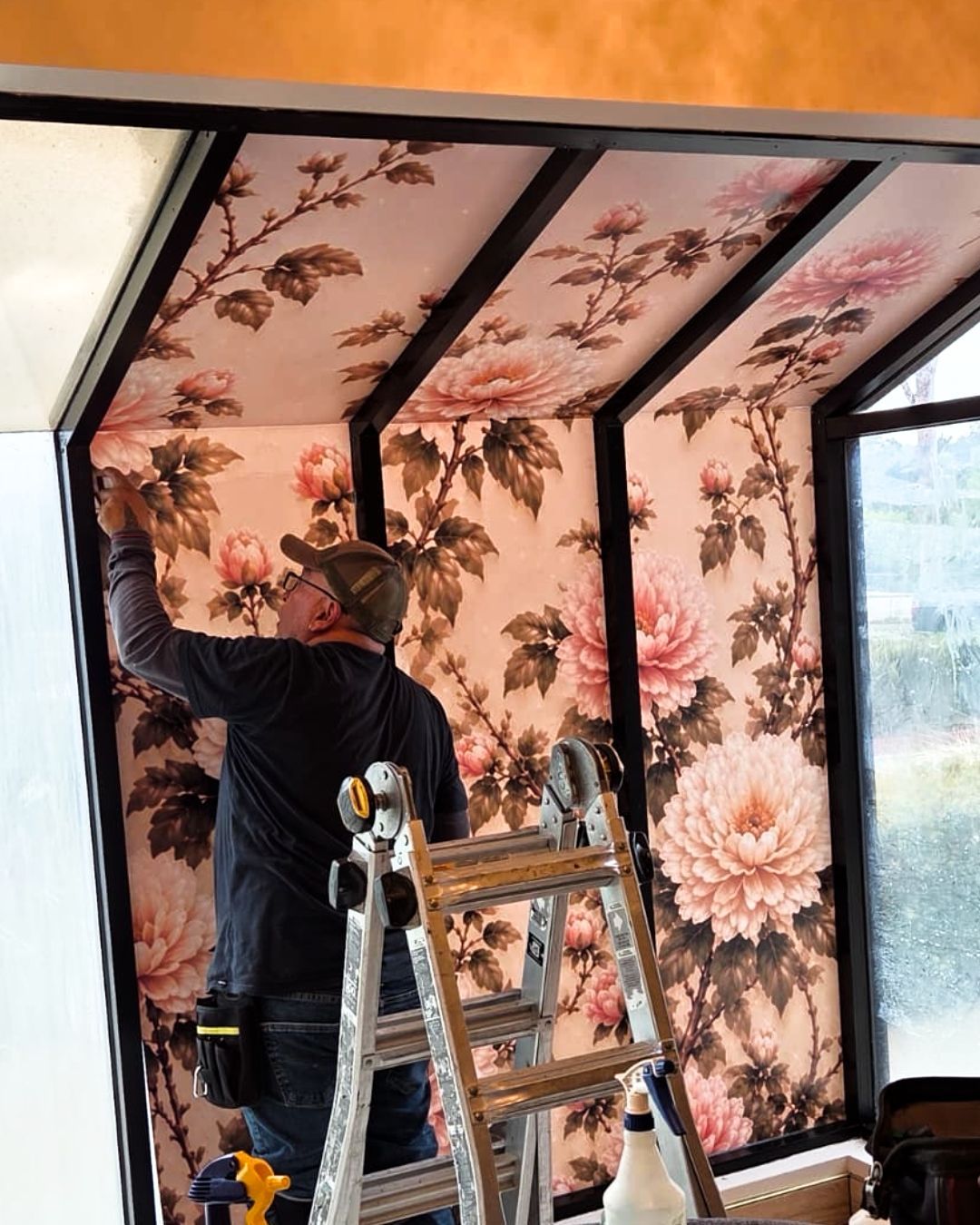 Person installing floral wallpaper on a sunroom wall beside a ladder.